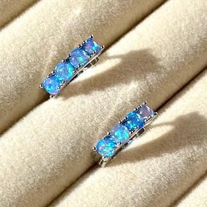 Peut inclure: Une paire de boucles d'oreilles argentées, chacune ornée de cinq opales ovales bleu clair. Les opales sont disposées en ligne, créant un design linéaire. Les boucles d'oreilles sont présentées sur une surface beige texturée.