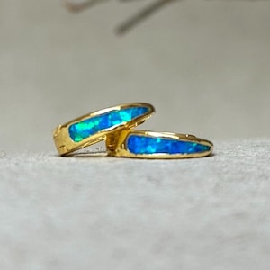 May include: A pair of gold hoop earrings featuring a blue opal inlay. The opal displays a shimmering, iridescent effect with green and blue hues. The earrings are presented on a neutral surface, accentuating their design and colour.