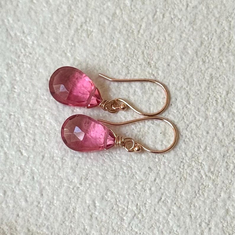 May include: A pair of pink teardrop-shaped earrings with faceted surfaces. The earrings are suspended from delicate, gold-colored ear wires. The gemstones have a translucent quality, and the overall design is elegant and simple.