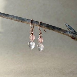 May include: Rose gold dangle earrings with a clear, teardrop-shaped crystal and a pink bead. The earrings are suspended from a delicate rose gold hook. The earrings are displayed on a small branch.