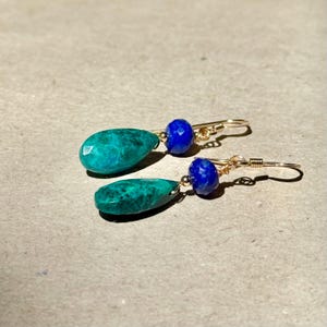 May include: A pair of gold earrings. Each earring has a teal, teardrop-shaped stone and a small, round, blue stone. The earrings are presented on a neutral-toned surface.