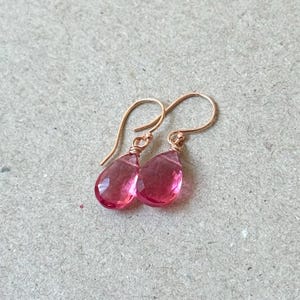 May include: A pair of rose gold earrings with teardrop-shaped, faceted pink gemstones. The earrings have a simple hook design and are displayed on a neutral background. The gemstones are a vibrant shade of pink.