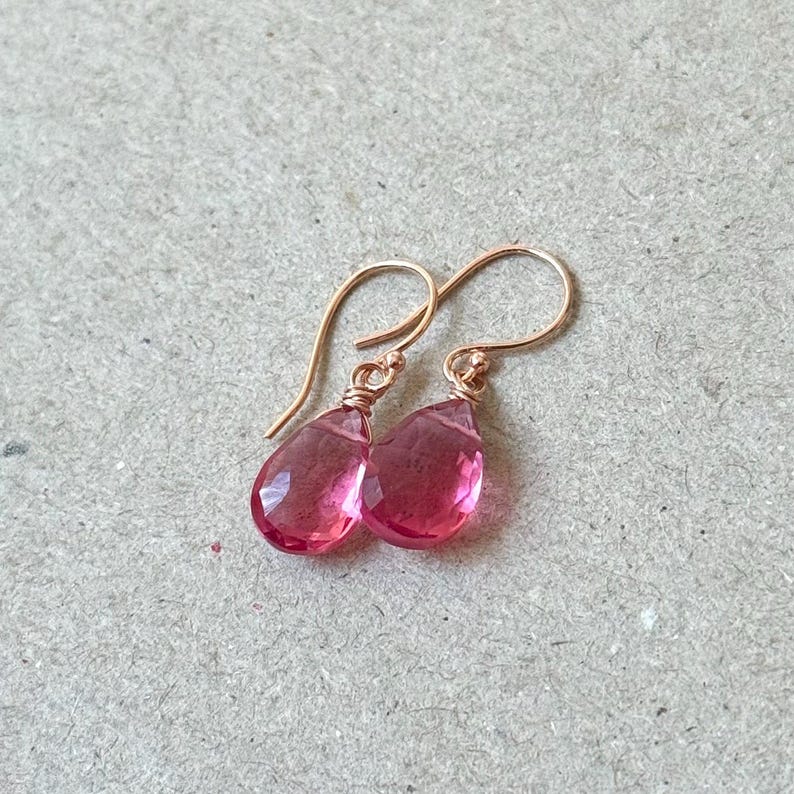 May include: A pair of rose gold earrings with teardrop-shaped, faceted pink gemstones. The earrings have a simple hook design and are displayed on a neutral background. The gemstones are a vibrant shade of pink.
