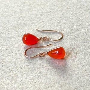 May include: A pair of handcrafted earrings featuring teardrop-shaped, vibrant orange gemstones. The gemstones are wire-wrapped and suspended from delicate gold-colored ear wires, creating a simple yet elegant design. The earrings are displayed on a neutral background.