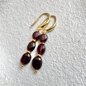 May include: A pair of dangle earrings with three dark purple oval beads on each earring. The beads are separated by gold spacers. The earrings have gold hooks.