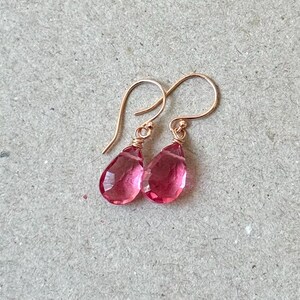 May include: A pair of rose gold earrings with teardrop-shaped, faceted pink gemstones. The gemstones are wire-wrapped and suspended from delicate ear wires, creating a simple yet elegant design. The earrings are displayed on a neutral background.