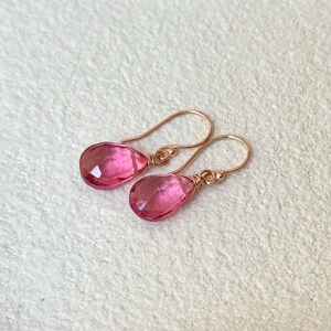 May include: A pair of pink teardrop-shaped earrings with rose gold ear wires. The faceted pink gemstones are wire-wrapped to the ear wires. The earrings are displayed on a white surface.