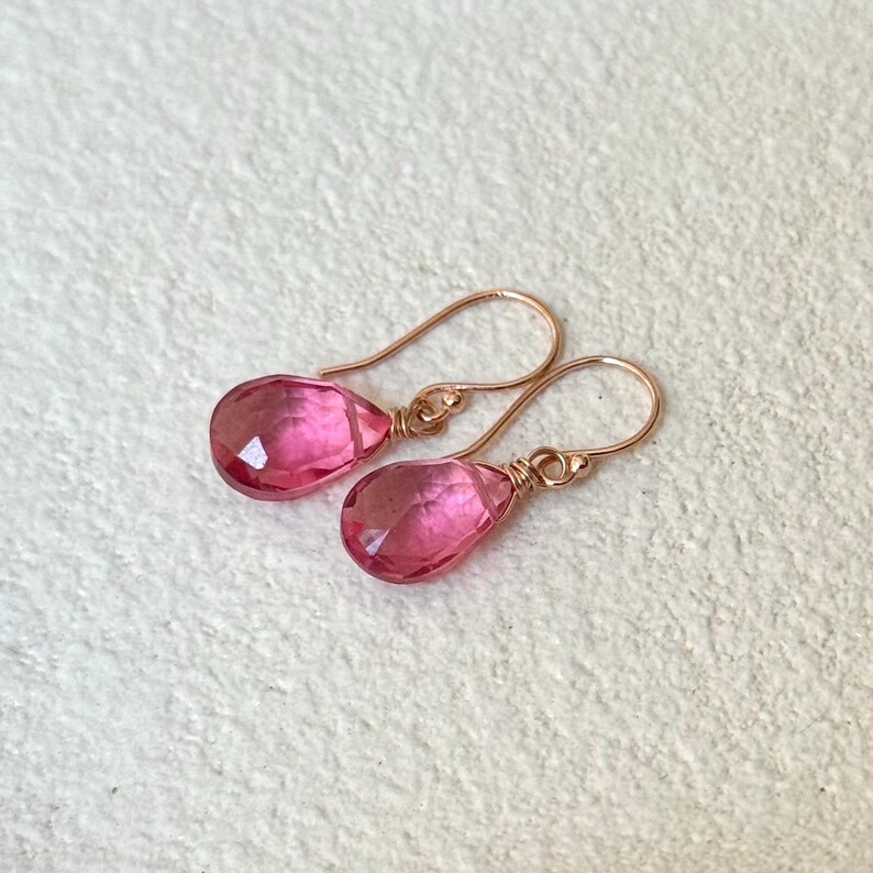 May include: A pair of pink teardrop-shaped earrings with rose gold ear wires. The faceted pink gemstones are wire-wrapped to the ear wires. The earrings are displayed on a white surface.