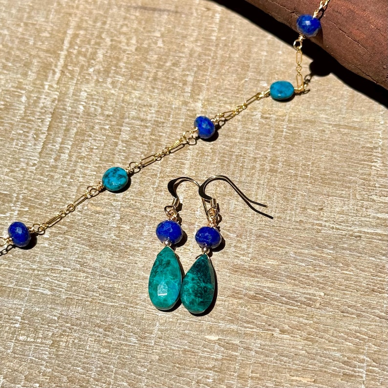 May include: A gold chain necklace and matching earrings set. The necklace and earrings feature blue and teal-coloured beads. The earrings have teardrop-shaped teal stones. The jewellery is displayed on a wooden surface.
