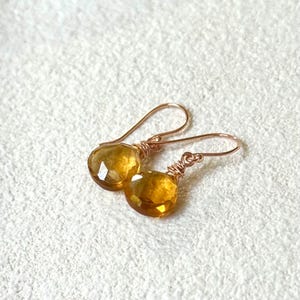 May include: A pair of gold-colored earrings with faceted, round, amber-colored gemstones. The gemstones are wire-wrapped and suspended from curved ear wires. The earrings are displayed on a textured, off-white surface.