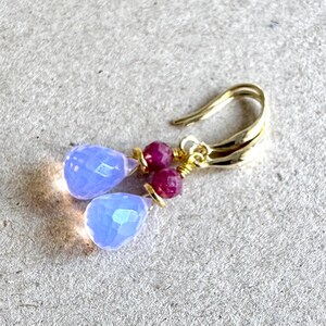 May include: A pair of gold dangle earrings featuring a teardrop-shaped light blue gemstone and a small red gemstone on each earring.
