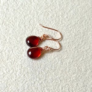 May include: A pair of teardrop-shaped earrings with deep red, translucent stones. The stones are wire-wrapped in a rose gold color, and the ear wires are also rose gold. The earrings are displayed on a textured, off-white surface.