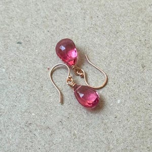 May include: A pair of rose gold earrings with faceted, teardrop-shaped pink gemstones. The earrings have a simple hook design and are displayed on a neutral-toned surface. The gemstones are a vibrant pink color.