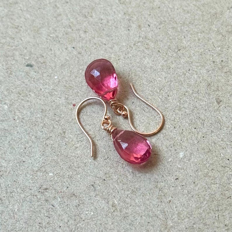 May include: A pair of rose gold earrings with faceted, teardrop-shaped pink gemstones. The earrings have a simple hook design and are displayed on a neutral-toned surface. The gemstones are a vibrant pink color.