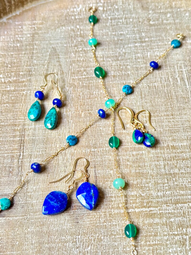 May include: A selection of gold-coloured jewellery, including necklaces and earrings. The necklaces and earrings are decorated with various gemstones in shades of blue and green. The earrings are teardrop-shaped.