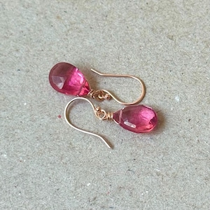 Puede incluir: Un par de pendientes colgantes con gemas rosas facetadas en forma de lágrima. Las gemas cuelgan de delicados ganchos dorados. Los pendientes se exhiben sobre una superficie neutra y texturizada.