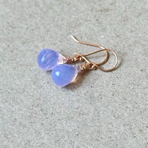 May include: A pair of delicate drop earrings featuring faceted, light purple teardrop-shaped gemstones. The gemstones are wire-wrapped and suspended from gold-colored ear wires. The earrings are displayed on a neutral-toned surface.