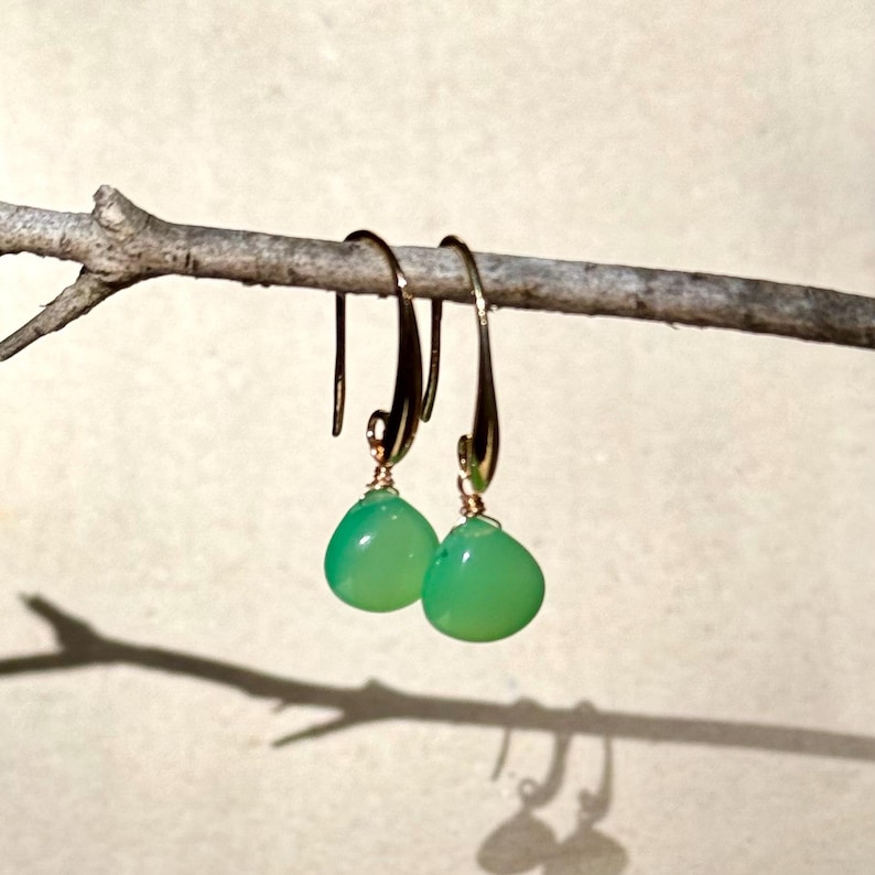 May include: A pair of green gemstone earrings with gold hooks. The earrings are hanging from a branch.