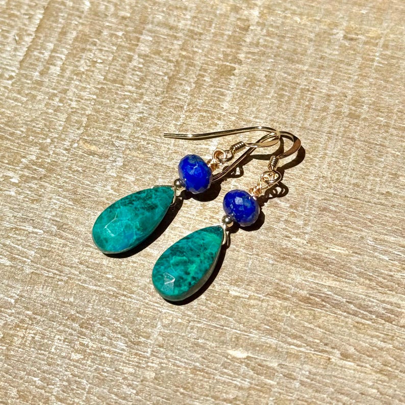 May include: A pair of gold earrings featuring a teal, teardrop-shaped stone and a small, round, blue stone. The earrings are presented on a light brown wooden surface.