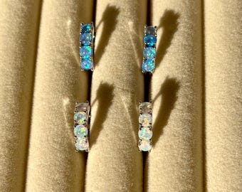 May include: Four pairs of silver-toned hoop earrings. Two pairs feature blue opal stones, and the other two pairs have white opal stones. The earrings are displayed on a beige surface.
