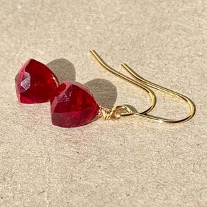 Puede incluir: Un par de pendientes de piedras preciosas de color rojo rubí. Cada pendiente presenta una piedra preciosa roja facetada, de forma triangular, suspendida de un gancho de alambre dorado. Las piedras miden aproximadamente 1,3 cm.