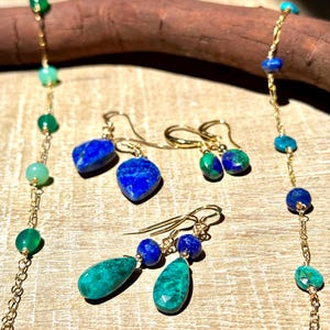 May include: A selection of gemstone jewellery, including necklaces and earrings. The jewellery features gold chains and various gemstones in shades of blue, green, and gold. The earrings have a drop design, and the necklaces have a beaded style.