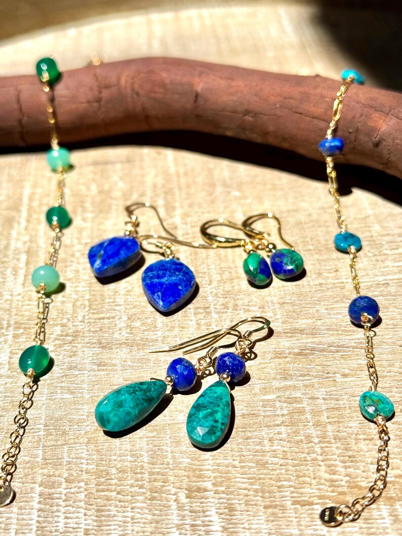 May include: A selection of gemstone jewellery, including necklaces and earrings. The jewellery features gold chains and various gemstones in shades of blue, green, and gold. The earrings have a drop design, and the necklaces have a beaded style.
