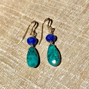 May include: A pair of gold earrings with a dark blue, round bead and a teal teardrop-shaped stone. The earrings are displayed on a light brown wooden surface.