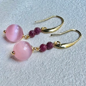 Puede incluir: Pendientes de oro con cuentas de piedras preciosas rosas y moradas. Los pendientes tienen un diseño colgante con una cuenta rosa más grande en la parte inferior.