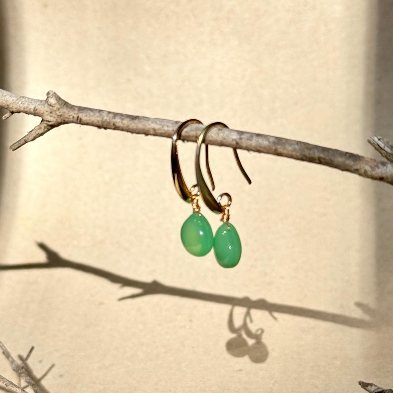 May include: A pair of gold hoop earrings with green gemstone drops. The earrings are hanging from a branch.