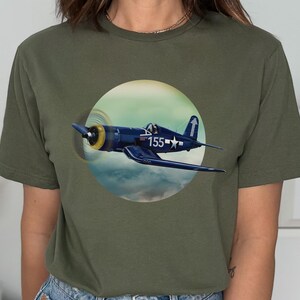 May include: A green t-shirt with a navy blue airplane graphic. The airplane is a Vought F4U Corsair with the number 155 on the side. The airplane is flying in a cloudy sky.