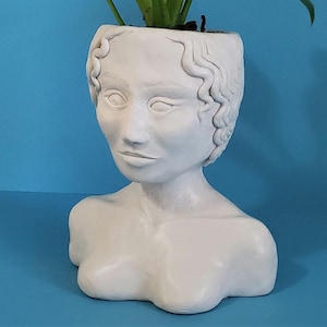 May include: White ceramic planter shaped like a bust of a woman with wavy hair. The planter is empty.