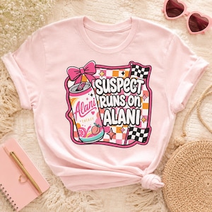 May include: A light pink t-shirt featuring a graphic design with the text "SUSPECT RUNS ON ALANI". The design includes a pink bow, a can of "Alani Energy", and checkered flags. The shirt is a crew neck style.