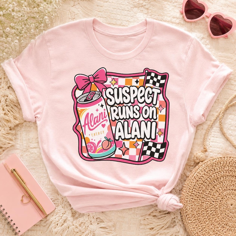 May include: A light pink t-shirt featuring a graphic design with the text "SUSPECT RUNS ON ALANI". The design includes a pink bow, a can of "Alani Energy", and checkered flags. The shirt is a crew neck style.