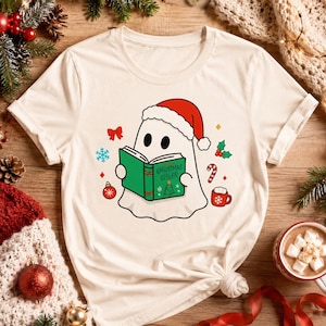 K&ouml;nnte beinhalten: Cremefarbenes T-Shirt mit einem Cartoon-Geist, der einen Weihnachtsmannhut tr&auml;gt und ein gr&uuml;nes Buch mit den Worten "Christmas Cheer" liest. Festliche Weihnachts-Grafiken, darunter eine Zuckerstange und eine Verzierung, umgeben den Geist.
