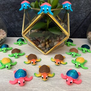 May include: A collection of small, colorful turtle figurines. The turtles are made of plastic and have different colors, including green, blue, pink, and brown. Some of the turtles are sitting on a wooden surface, while others are perched on a glass vase with a green plant.