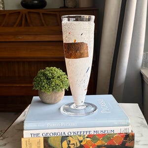 May include: A clear glass vase with a flared base and a textured white surface. The vase has a brown rectangular design and colourful speckles. It is displayed on a stack of books, including one titled "Pablo Picasso."