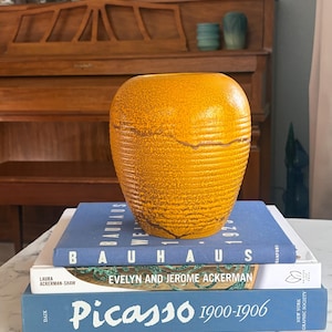 May include: A mustard yellow ceramic vase with a ribbed texture and a speckled finish, resting on a stack of three books titled Bauhaus, Evelyn and Jerome Ackerman, and Picasso 1900-1906, featuring blue and white covers.