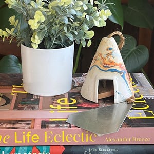 May include: A small, white ceramic teepee with a brown rope hanging from the top. The teepee has a colorful, abstract design with orange, blue, and white paint. The teepee is sitting on a stack of books with the title "Life Eclectic" visible.