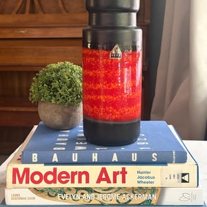 May include: A cylindrical ceramic vase with a matte black top and base, featuring a vibrant red and orange speckled midsection. A small triangular sticker with the text Gertz is visible on the front of the vase, which is resting on a stack of books.