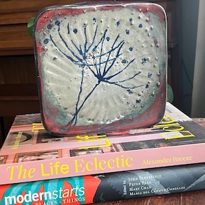 May include: A square ceramic tile with a hand-painted design of a stylized flower in shades of blue, white, and red. The tile has a textured surface and is sitting on top of two books.