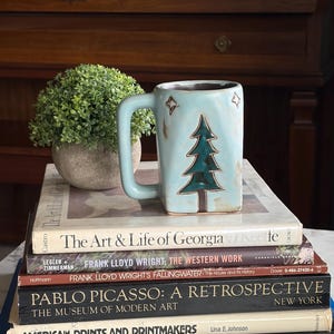 May include: A light blue ceramic mug with a Christmas tree design and star accents, resting on a stack of books about art and architecture. A small potted plant sits to the left of the mug. The mug has a square shape and a large handle.