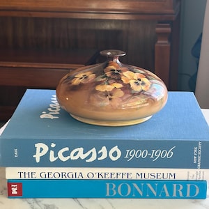 May include: A brown, oval-shaped vase with a narrow neck, adorned with painted yellow and orange floral designs. The vase rests atop a stack of books, including one titled "Picasso 1900-1906".