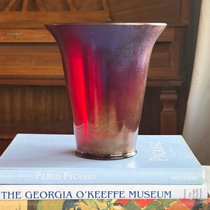 May include: A tall, flared vase with a gradient of red and purple hues. The glass vessel sits atop a stack of books, including one titled "Pablo Picasso". The vase's rim has a subtle gold tone.