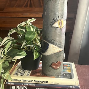 May include: A tall, cylindrical ceramic vase in shades of gray, adorned with stylized eyes and a mouth. The vase sits atop a stack of books, including "American Prints and Printmakers" and "The Fauves". A potted plant is to the left.