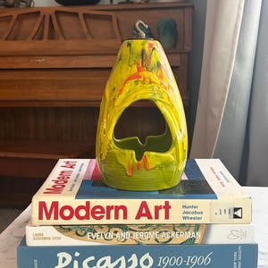 Peut inclure: Un objet en céramique jaune et vert avec un vernis dégoulinant, une ouverture centrale et un crochet métallique au sommet, posé sur une pile de trois livres intitulés Modern Art, Evelyn and Jerome Ackerman et Picasso 1900-1906.