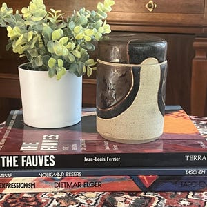 Peut inclure: Un pot en céramique cylindrique avec un couvercle, orné d'un motif abstrait marron et beige. Il repose sur une pile de livres, dont "Les Fauves" et "Expressionnisme". Un petit pot blanc avec du feuillage vert est à gauche.
