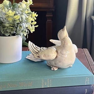 May include: A white ceramic bird figurine with brown accents, perched on top of two books. The top book is teal blue and has the text "American Art Deco 1925-1939" printed on the spine. The bottom book is dark blue and has the text "Whitney" printed on the spine.