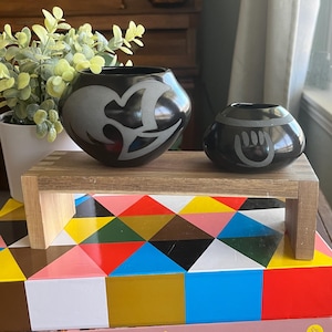 May include: Two black pottery bowls with white geometric designs. The bowls are sitting on a wooden stand. The stand is on top of a stack of books with colorful geometric patterns.
