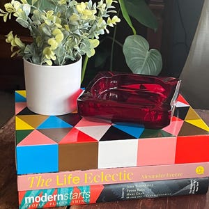 May include: A red glass ashtray with a geometric pattern on a stack of books. The top book is titled "The Life Eclectic" and the bottom book is titled "modernstarts PEOPLE PLACES THINGS".